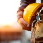 Close-up of a construction worker holding a yellow hard hat, with a tool belt equipped with a hammer and other tools, against a blurred background of a job site with warm sunlight.